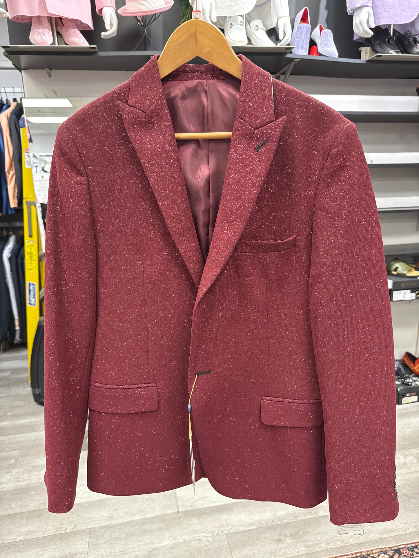 The EJ SAMUEL JACKET 19 CLEARANCE, featuring a maroon hue and notched lapel, is displayed on a wooden hanger in a clothing store with shelves and other garments visible in the background; please note this item is final sale and cannot be returned or exchanged.
