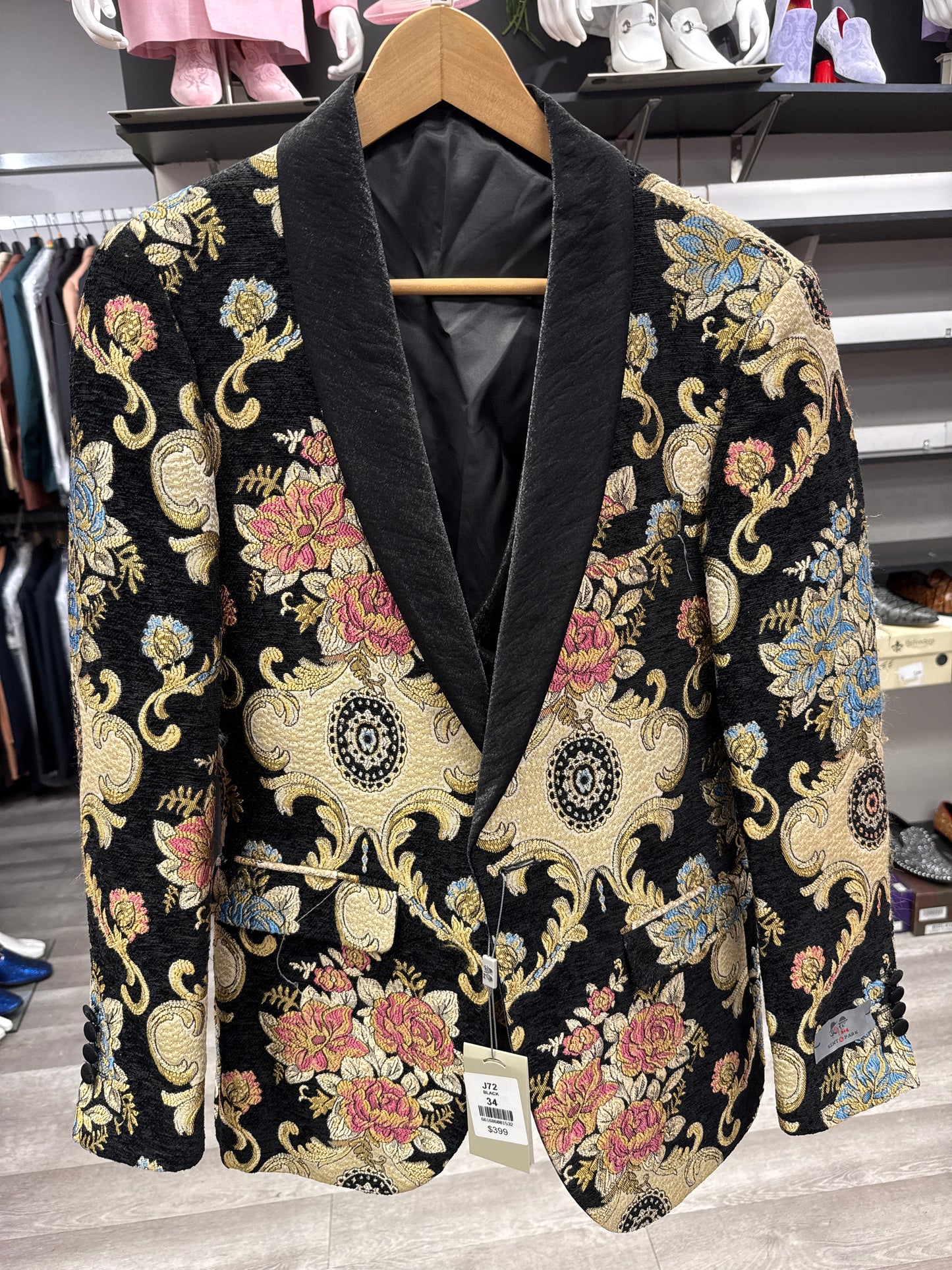 JACKET 13 CLEARANCE by EJ SAMUEL: Black blazer with a floral brocade pattern in gold, blue, red, and tan tones, featuring a shawl collar and displayed on a wooden hanger in a clothing store. Please note: this item is final sale with no returns or exchanges.