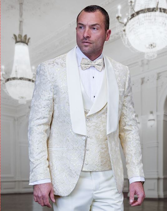 A man in a STATEMENT TIZANO OFF WHITE Suit by Statement Clothing, featuring a matching bow tie and vest, stands elegantly in a room adorned with chandeliers.