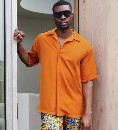 A man wearing the Inserch 2pc Set (Shirts and Pants) in Burnt Orange by Inserch stands by a doorway, holding the door frame with one hand and wearing sunglasses.