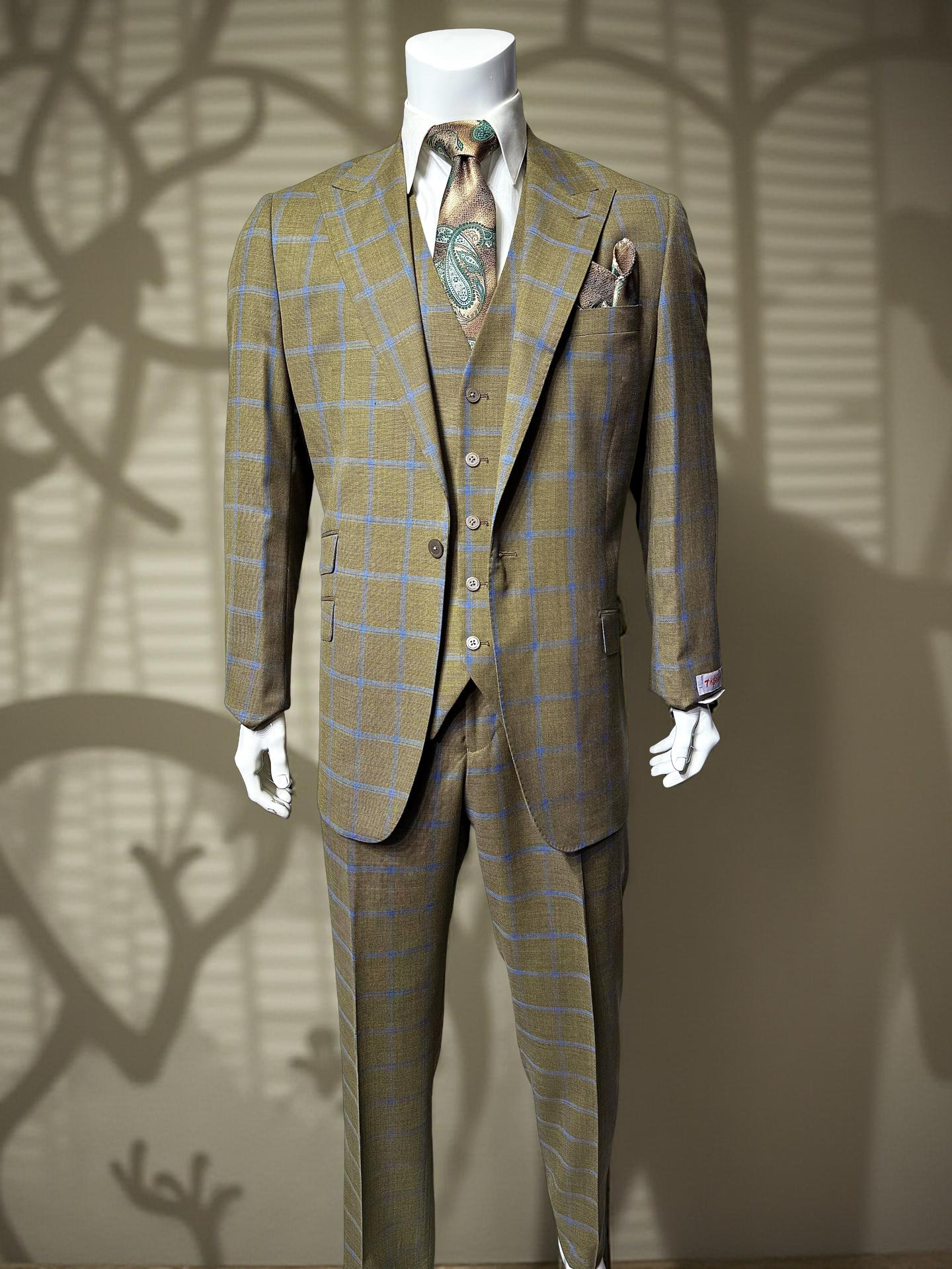 A tailored mannequin showcases the Tiglio Rosso Portofino Semi Wide Leg TL8068 suit in tan checkered 100% wool with matching vest, tie, and pocket square, set against a wall with abstract shadow patterns.