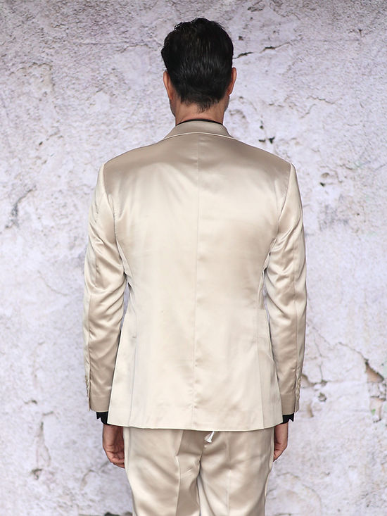 A man with short dark hair and a beard wears the Insomnia INSOMNIA CLASSICO 3PC Tailored Fit Satin Spandex Champagne Suit with a black shirt, standing before a textured light wall—ideal for a stylish night out.