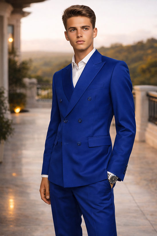 A young man stands on a terrace, wearing the Manzini Impress Men's 2PC Spandex Double Breast Slim-Fit Suit in royal blue and a white shirt, with scenic trees and hills in the background.