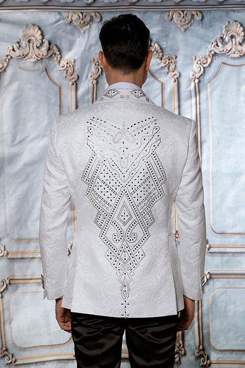 A man wears the Manzini JACKET 37 CLEARANCE—a double-breasted, patterned white tuxedo jacket with metallic lapels and a bow tie—standing before an ornate, light blue and gold decorative backdrop. The jacket’s subtle floral textured pattern enhances the depth and sophistication of his ensemble.