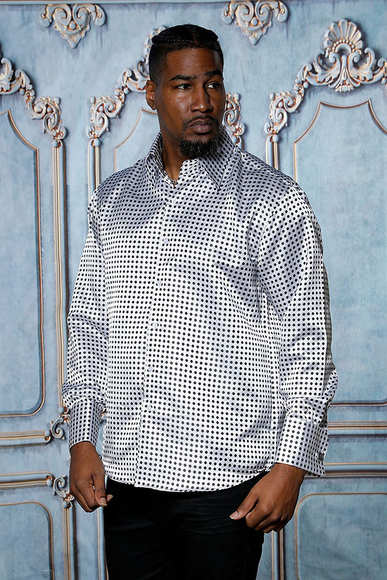 A man wearing the Manzini MANZINI MZT-164 Small Polka-Dot Long Sleeve Satin Shirt with cuff and link in white stands before an ornate, light blue decorative wall.