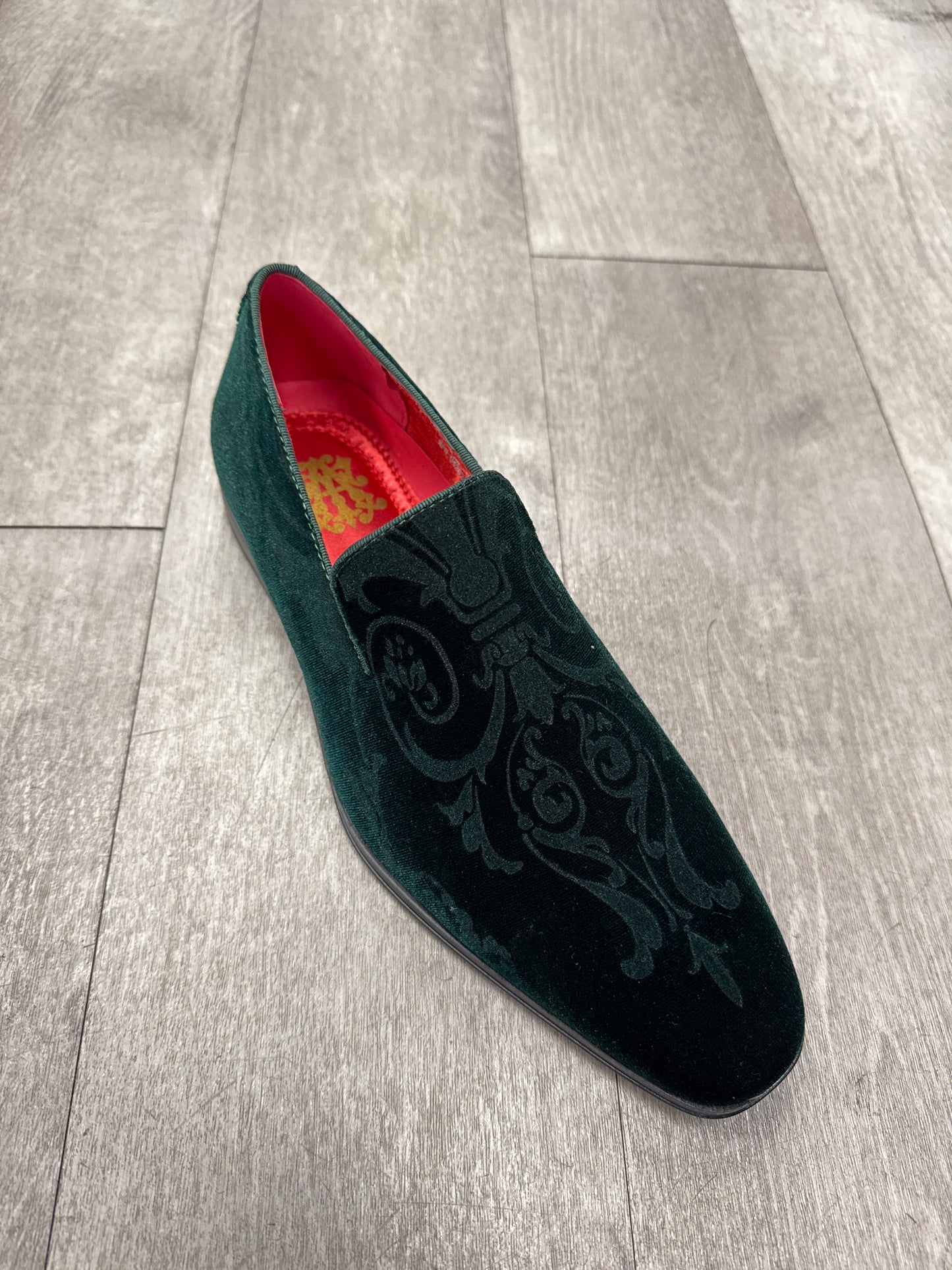 The After Midnight 7017 Green Paisley loafer from AFTER MIDNIGHT, featuring the ornate embroidery and a sleek buckle accent, rests on a wooden floor. Its interior is lined with rich red fabric.