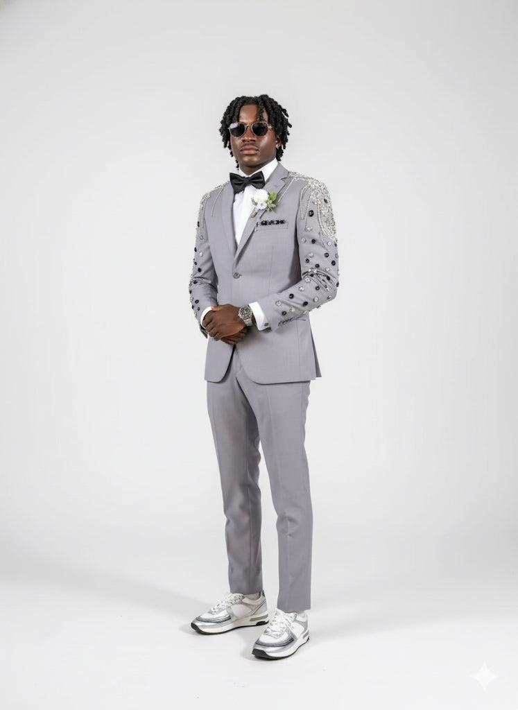 A man in the Unique Design Menswear Custom Prom Suit Grey with Black/Silver Stone Work and metallic sneakers poses against a white background, accessorizing with a bow tie and sunglasses.