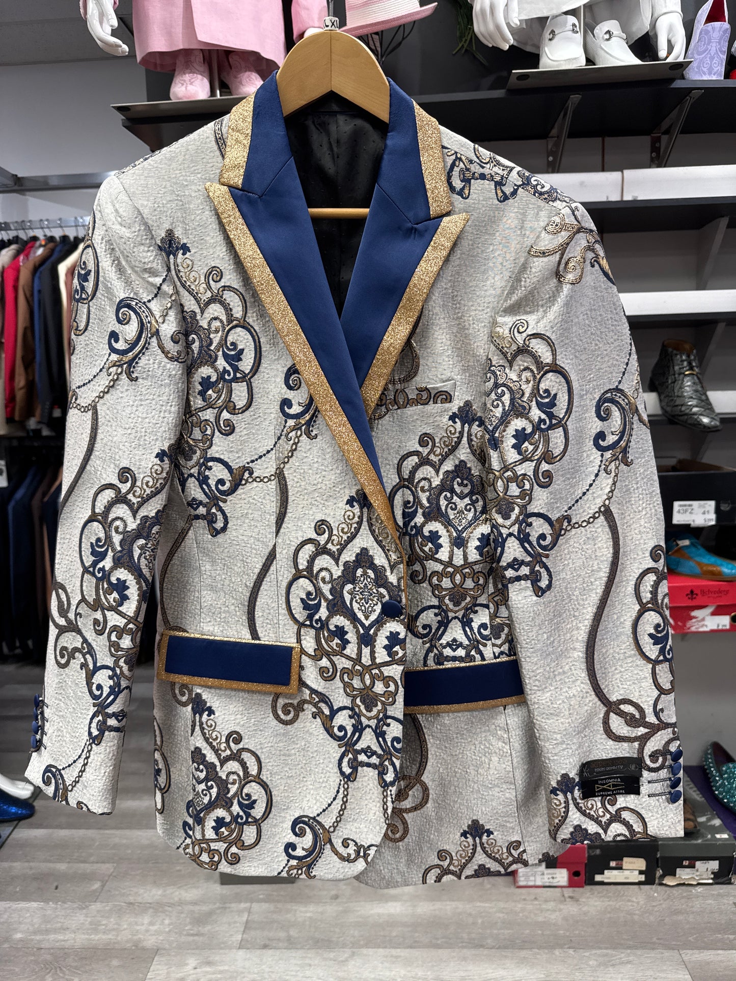 The EJ SAMUEL JACKET 8 CLEARANCE, featuring a patterned cream and blue design with gold trim, is displayed on a hanger in a clothing store; please note that this is a final sale item with no exchanges or returns.