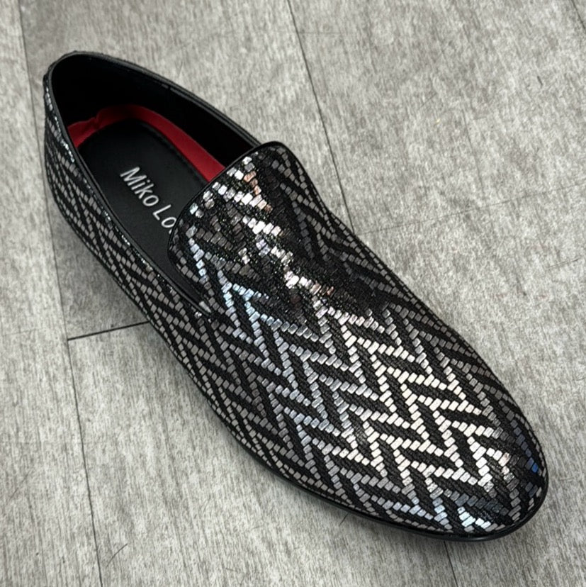 The Exclusive Formal Dress Shoe Silver / Black B2383 by UNIQUE DESIGN MENSWEAR, featuring a zigzag pattern crafted from genuine leather with black and silver sequins, is placed on a wooden floor.