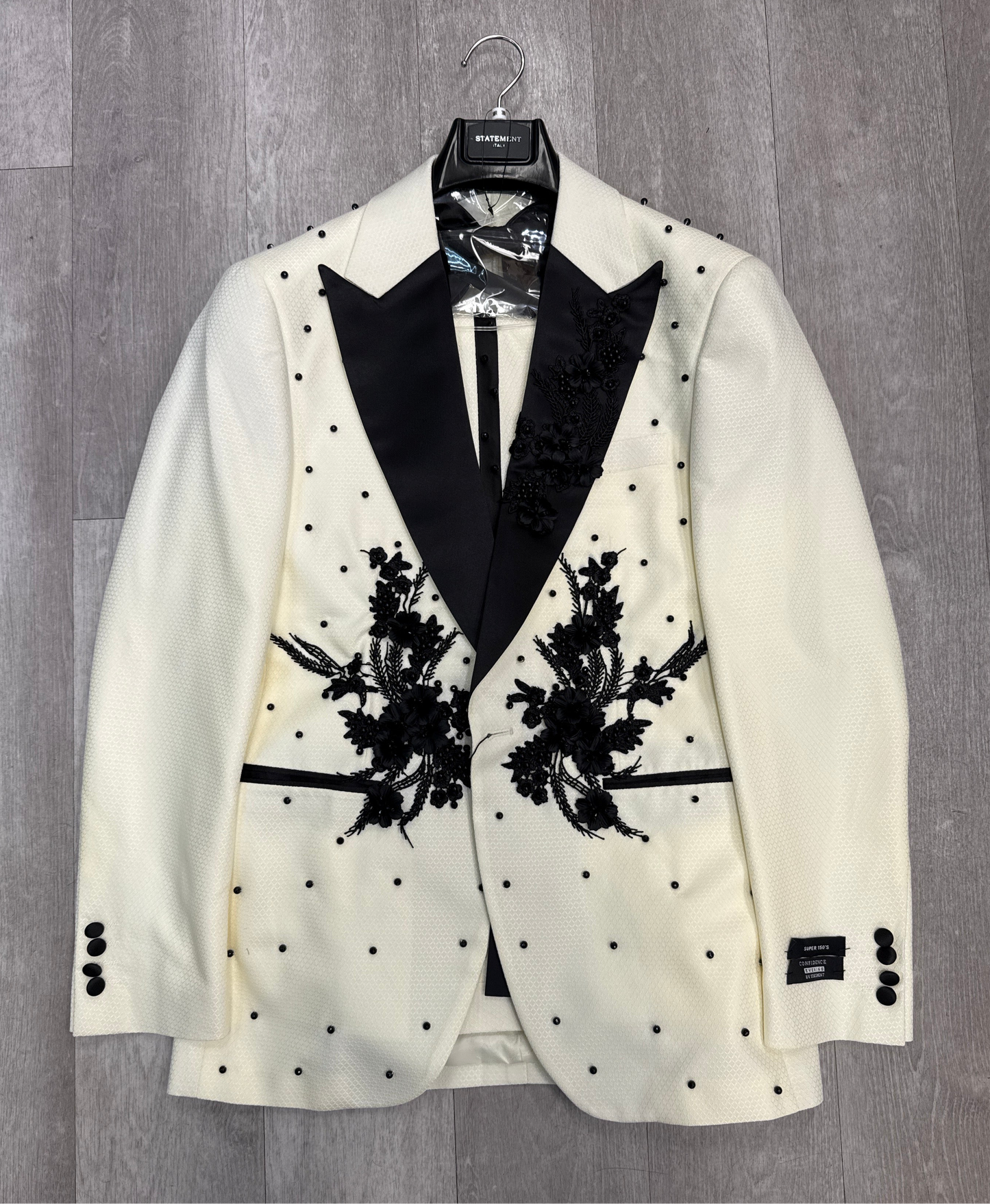 The STATEMENT CLOTHING | PERINI - OFFWHITE - BLACK tuxedo jacket by Statement Clothing features a cream color, black lapels, black floral embroidery, and black polka dot accents, shown on a hanger against a gray wood floor.
