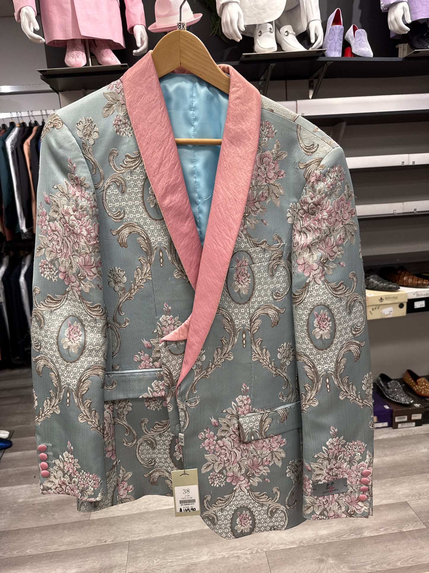The EJ SAMUEL JACKET 20 CLEARANCE, a men's blazer with a silver and pink floral brocade pattern on light blue fabric and a pink shawl collar, is displayed on a wooden hanger in the clothing store. Please note: this item is final sale—no exchanges or returns.