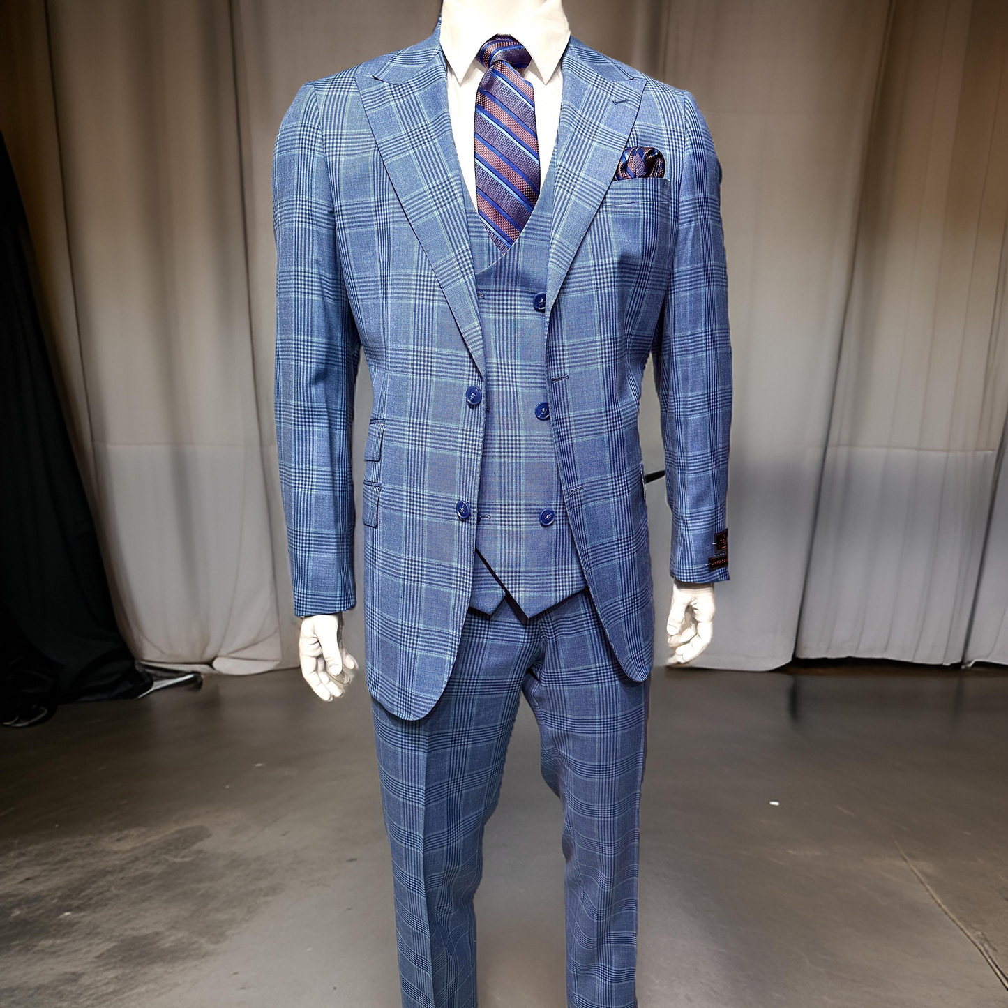 A mannequin wears the Unique Design Menswear Tiglio Luxe Prosecco Modern Fit Suit/Vest Blue Plaid TL5587, styled with a matching vest, striped tie, and pocket square on a polished floor before neutral curtains—classic menswear showcased.