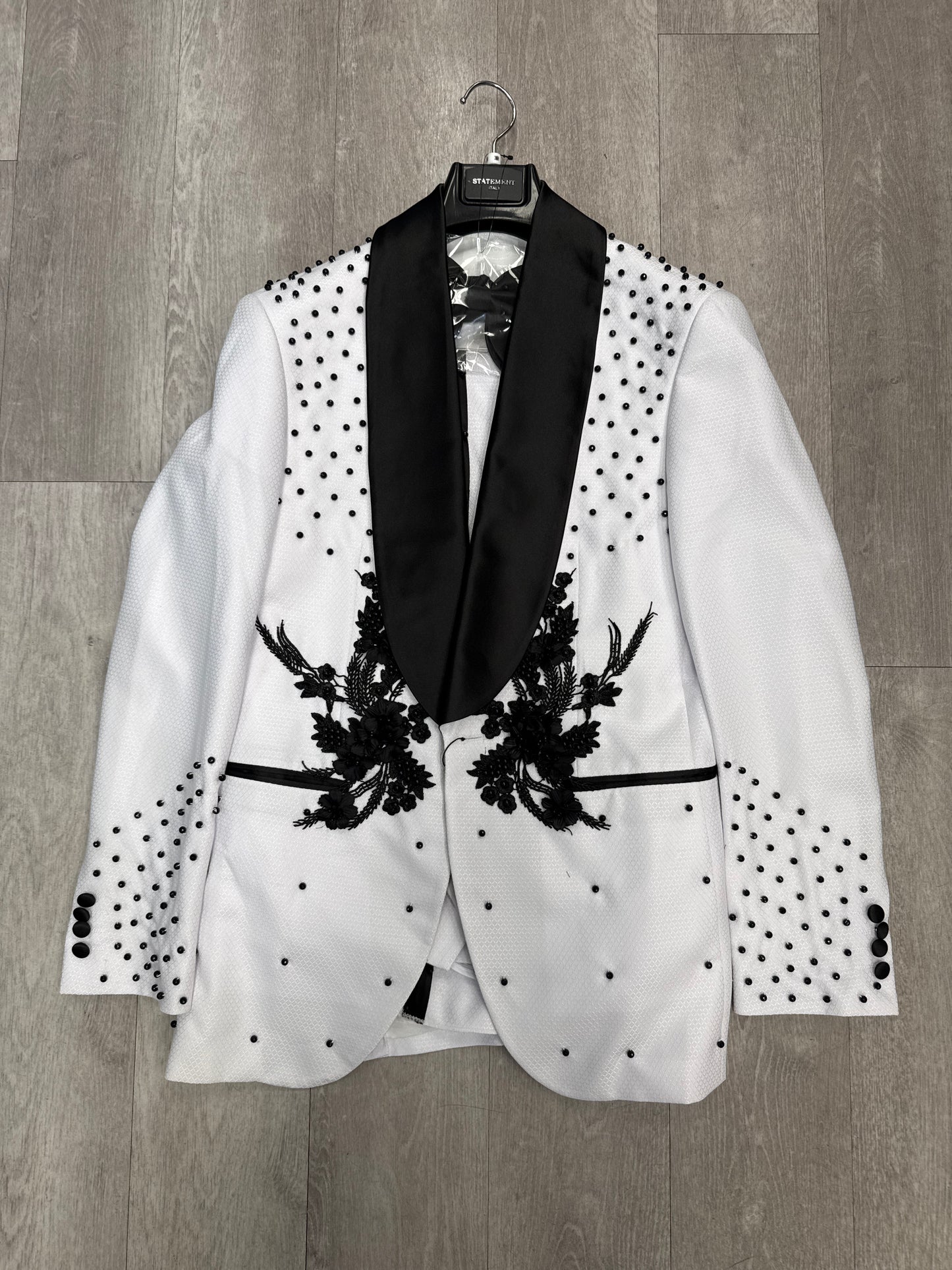 The STATEMENT CLOTHING | ELIO-WHITE WITH BLACK STONES tuxedo blazer by Statement Clothing features black floral embroidery, black lapels, and scattered black studs, displayed on a hanger against a wooden floor.