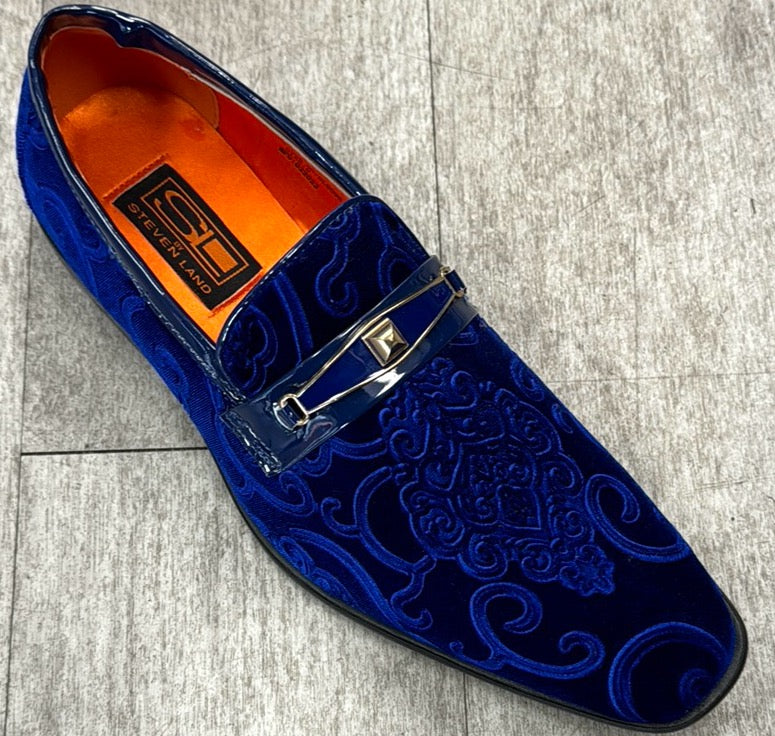 The "Exclusive Formal Dress Shoe Blue Paisley with Buckle SL0096" by UNIQUE DESIGN MENSWEAR features a blue velvet exterior adorned with intricate swirl patterns, complemented by a metallic buckle and an orange interior, all set against a gray floor and crafted from genuine suede leather.