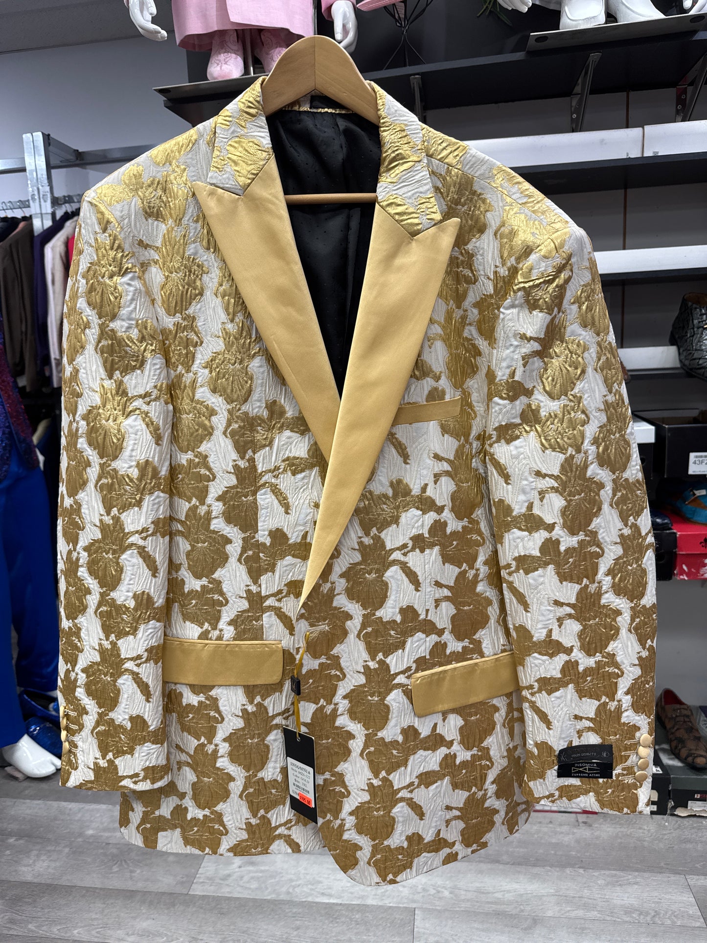 The EJ SAMUEL JACKET 4 CLEARANCE, featuring a gold and cream floral pattern with solid gold lapels and pockets, hangs on a wooden hanger in a clothing store. Please note: this item is final sale—no exchanges or returns permitted.