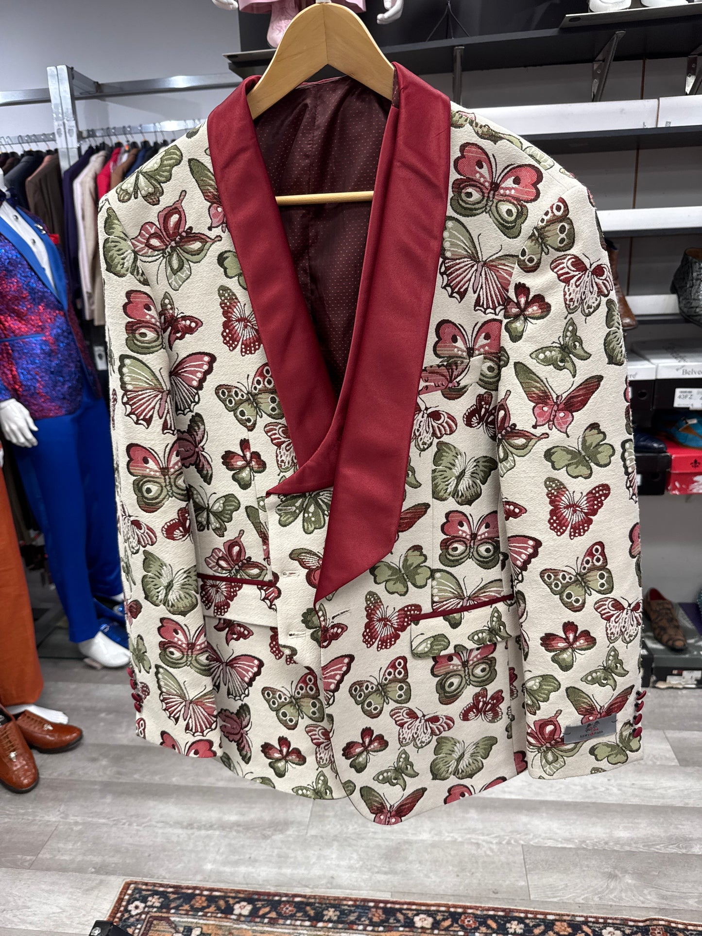 A cream EJ SAMUEL JACKET 26 CLEARANCE with red lapels and a green and red all-over butterfly print hangs on a wooden hanger in a clothing store. Please note: this item is a final sale.