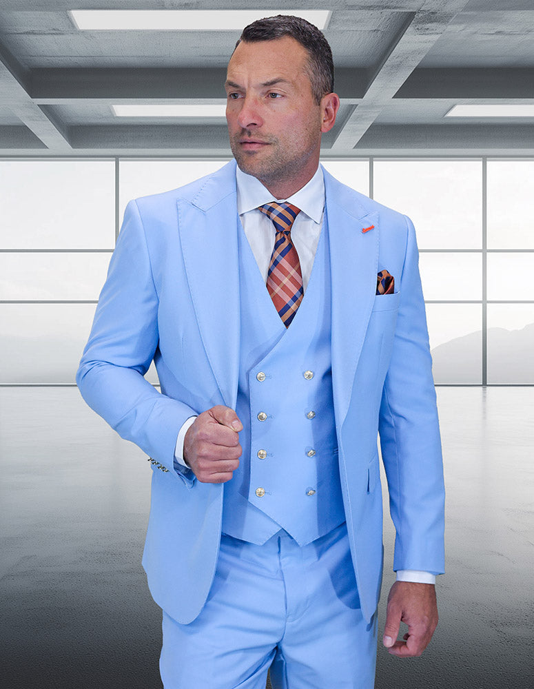 A man wearing the STATEMENT CLOTHING | AMIRI-SKY BLUE suit by Statement Clothing, paired with a white shirt and plaid tie, stands in a modern empty room with large windows.