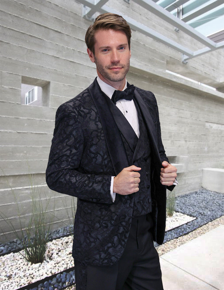 A man in the Statement Clothing AMORE-BLACK modern fit lace tuxedo with 100% wool pants and a bow tie stands before a sleek concrete building surrounded by rocks and plants.