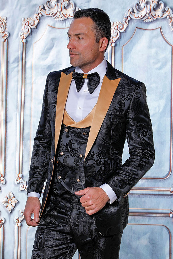 A man wears the MANZINI ARMANI 3-Piece Slim Fit Satin Filigree Pattern Black/Gold Tuxedo Prom Suit by Manzini, featuring gold lapels, a matching vest, and a large black bow tie, standing before an ornate light blue and white wall.