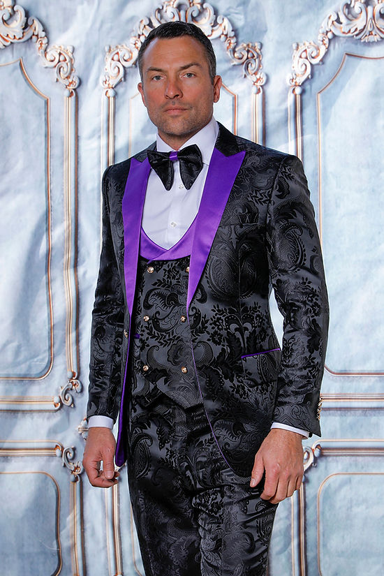A man in a Manzini MANZINI ARMANI 3-Piece Slim Fit Satin Filigree Pattern Black/Purple Tuxedo Prom Suit stands before an ornate, light blue and white backdrop.