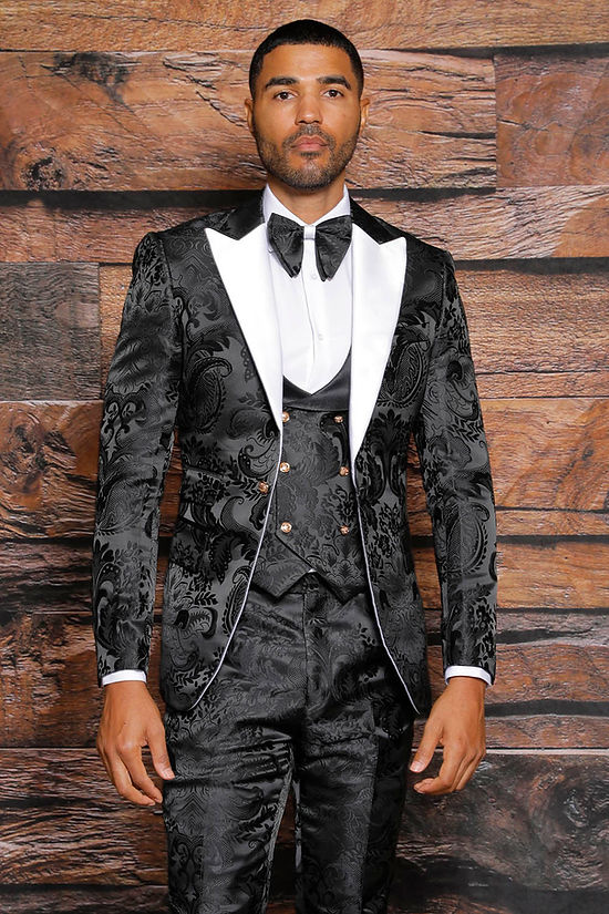 A man wears a Manzini Armani 3-Piece Slim Fit Satin Filigree Pattern Black/White Tuxedo Prom Suit with a white dress shirt and black bow tie, standing before a wooden background.