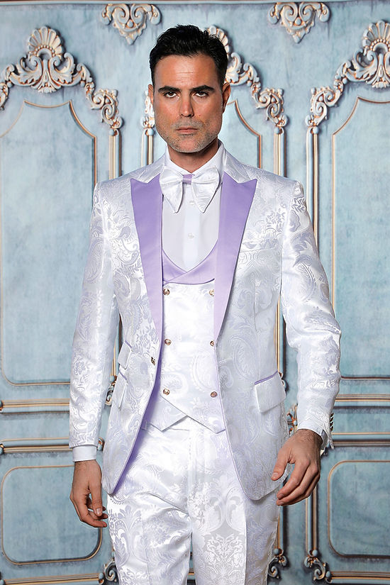 A man wears a Manzini ARMANI 3-piece slim fit satin filigree pattern white/lavender tuxedo prom suit, featuring lavender lapels, vest, and bow tie, standing against an ornate pale blue background.
