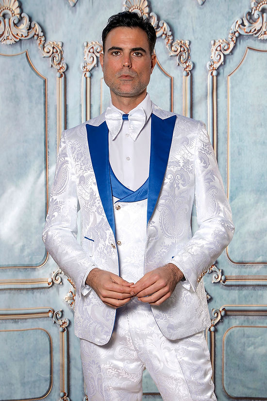 A man stands before a decorative wall in the Manzini Armani 3-Piece Slim Fit Satin Filigree Pattern White/Royal Tuxedo Prom Suit by Manzini, featuring a royal blue lapel, matching vest, and a white bow tie.