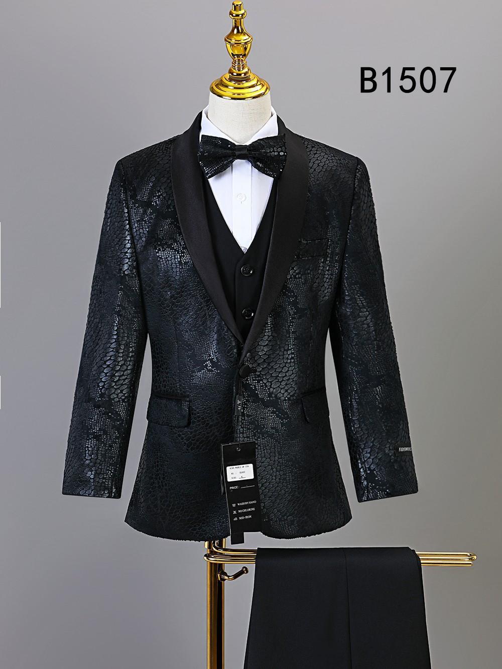 The BOY SUIT 5-PC SET B1507 by Unique Design Menswear features a black textured formal outfit with satin lapels, bow tie, vest, and pants displayed on a gold-stand mannequin—perfect for special occasions. Label tags and code B1507 visible.