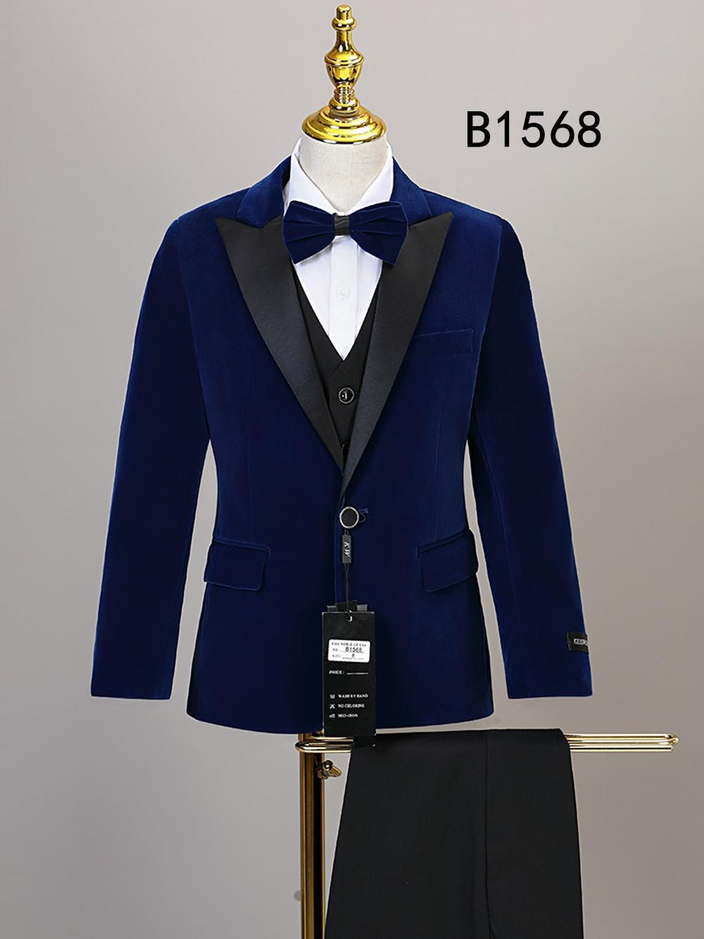 The BOY SUIT 5-PC SET B1568 by Unique Design Menswear includes a blue velvet tuxedo jacket with black lapels, white shirt, black waistcoat, bow tie, and black trousers—ideal formal wear for boys. "B1568" is shown on the wall behind the mannequin.