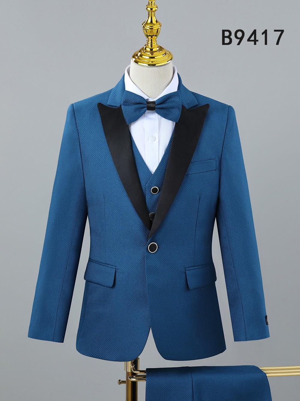 The BOY SUIT 5-PC SET B9417 by Unique Design Menswear features a blue suit with black lapels, white dress shirt, and blue bow tie—ideal for boys’ formal occasions. Displayed on a mannequin.