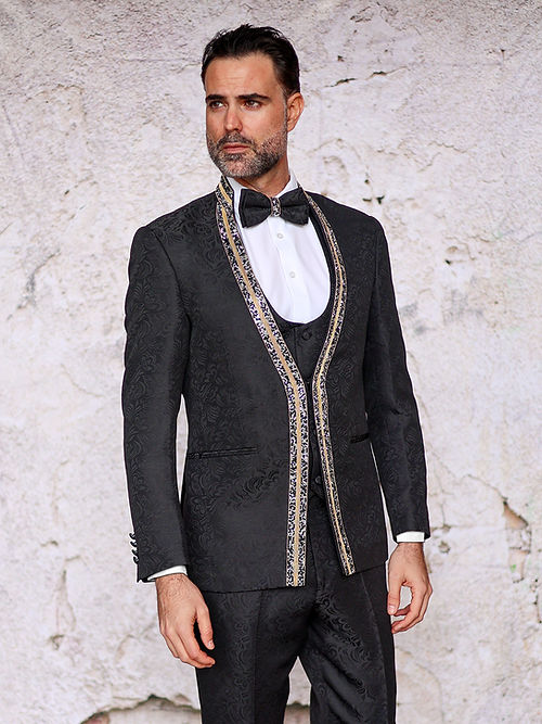 A man in a Manzini Beggio 3-Piece Slim Fit Glitter Trim Black Tuxedo Suit by Insomnia, featuring jacquard fabric and a bow tie, poses against a textured stone wall.