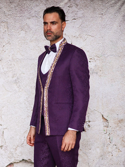 A man in an Insomnia MANZINI BEGGIO 3-piece slim fit eggplant tuxedo with glitter trim and a bow tie stands against a textured wall, the subtle floral pattern catching the light.