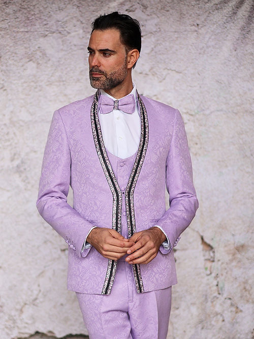 An elegant man stands against a textured wall in an Insomnia MANZINI BEGGIO lavender tuxedo suit, featuring glitter trim and floral patterns. The ensemble includes a white shirt and matching bow tie, highlighting the slim fit's sophistication, best maintained by dry cleaning only.