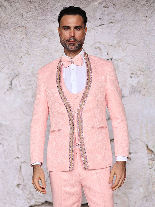 A man in the Insomnia MANZINI BEGGIO 3-Piece Slim Fit Glitter Trim Pink Tuxedo Suit stands poised against a light-textured wall, wearing a white shirt and pink bow tie, highlighting the slim fit and subtle floral pattern of his attire.