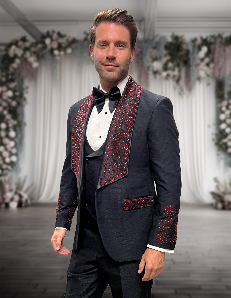 A man wears the STATEMENT CLOTHING | BELLA-BLACK tuxedo by Statement Clothing, featuring red embellishments and subtle rhinestones, standing in a formal venue decorated with flowers.