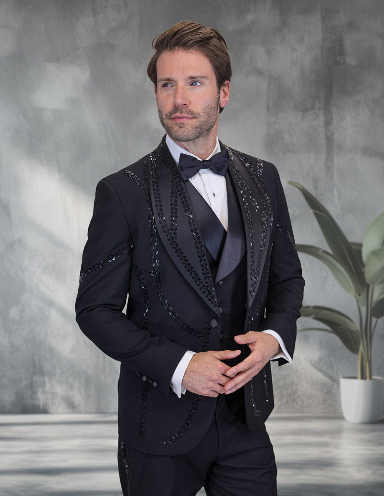 A man in the STATEMENT CLOTHING | CHIC-BLACK tuxedo by Statement Clothing stands indoors before a gray wall and potted plant, exuding modern black chic sophistication.