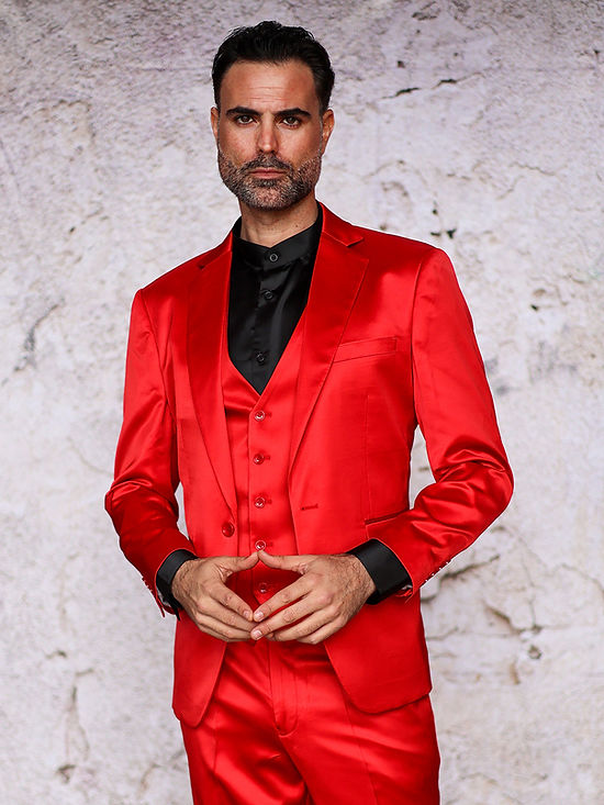 A man in the Insomnia INSOMNIA CLASSICO 3PC Tailored Fit Satin Spandex Red Suit with a black shirt stands against a textured light wall, showcasing the brand's ideal night-out style.