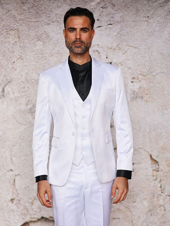 A man stands against a light-textured wall wearing the INSOMNIA CLASSICO 3PC Tailored Fit Satin Spandex White Suit by Insomnia, paired with a black dress shirt—ideal for a sophisticated night out.