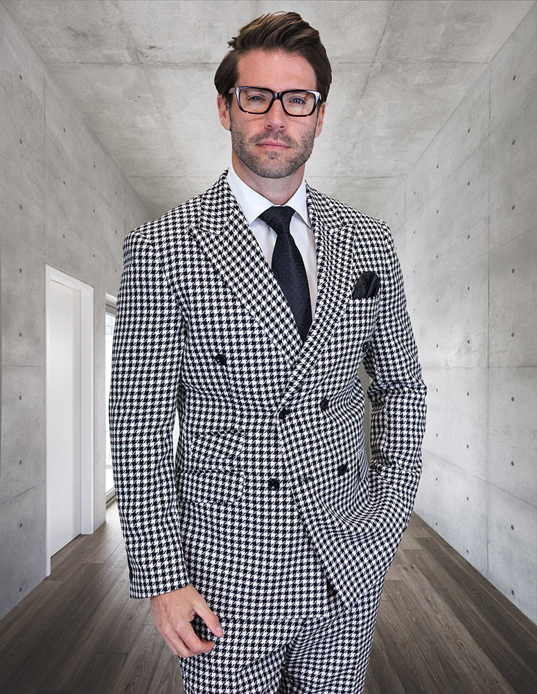 A man in a tailored-fit STATEMENT CLOTHING | DB MASIMO-BLACK houndstooth suit by Statement Clothing, white shirt, and black tie stands in a minimalist hallway with concrete walls and wooden floor.