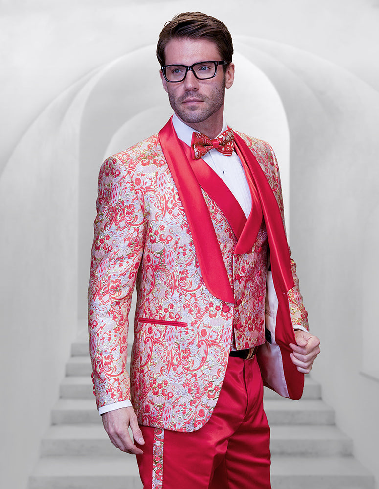 Wearing the STATEMENT CLOTHING | DELANO-2-CORAL by Statement Clothing, a man in glasses and a modern fit red and white patterned shawl lapel tuxedo with matching bow tie stands before white arched steps, radiating sophistication.
