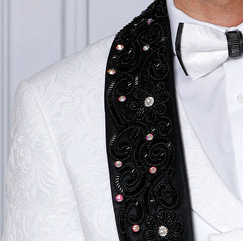Against a light wall, a confident man stands in an Insomnia MANZINI DIOR 3-piece slim fit rhinestone & bead trim floral white tuxedo suit with a black embellished scarf.