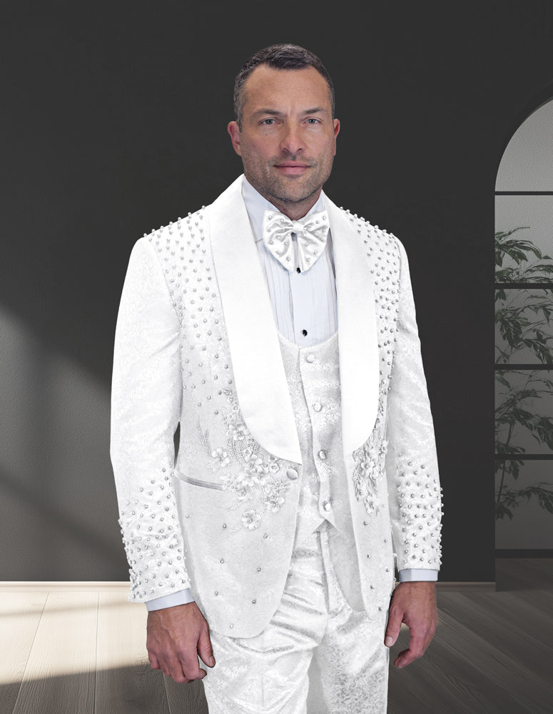 A man stands indoors wearing the STATEMENT CLOTHING | ELIO-WHT-WHT by Statement Clothing, a modern fit white tuxedo suit with decorative embellishments, bow tie, vest, and rhinestone-studded jacket.