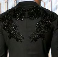 A man in an Insomnia MANZINI GQ300 Slim Fit Rhinestone Pattern Double Breast Black Tuxedo Suit, paired with a floral bow tie, stands against an ornate background.