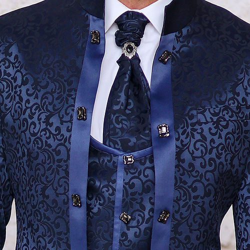 A man stands against a textured light wall wearing the MANZINI HAVANA 3-Piece Slim Fit Shiny Satin Pattern Banded Collar Sapphire Suit by Insomnia, featuring a fitted dark blue ensemble with a subtle floral pattern, matching vest, and tie. Dry clean only to maintain its pristine condition.