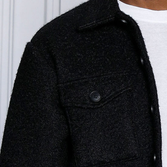 A man stands by a decorative white wall, gazing right, wearing an Insomnia MANZINI ICELAND Black Soft Fleece Button Sweater Shirt over a white shirt. This super soft poly fleece piece adds effortless style to his look.