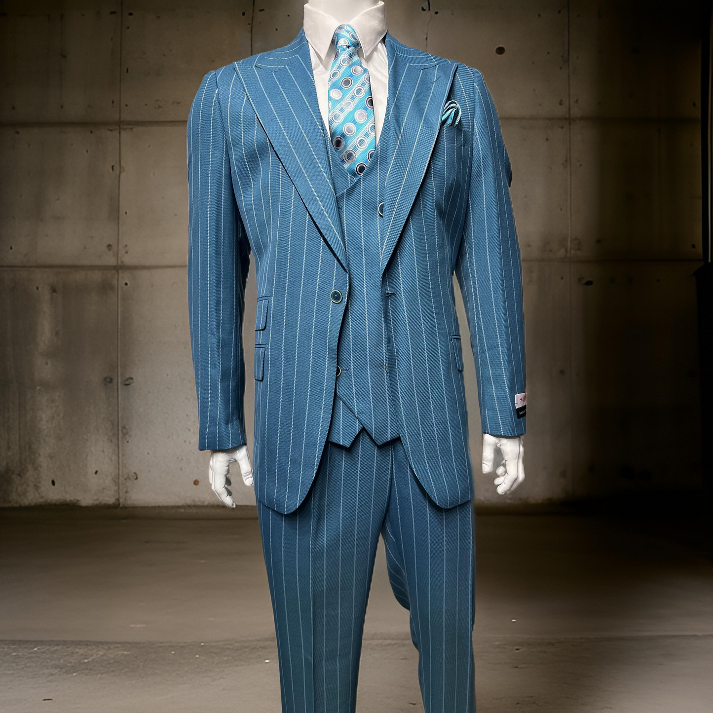 A man faces the camera in a Tiglio Rosso Orvietto Teal Blue Grey Pinstripe suit (TL6019) with wide-leg fit, white shirt, and patterned tie, standing against a plain light-colored wall. Available in sizes 42R, 48R, and 52R.
