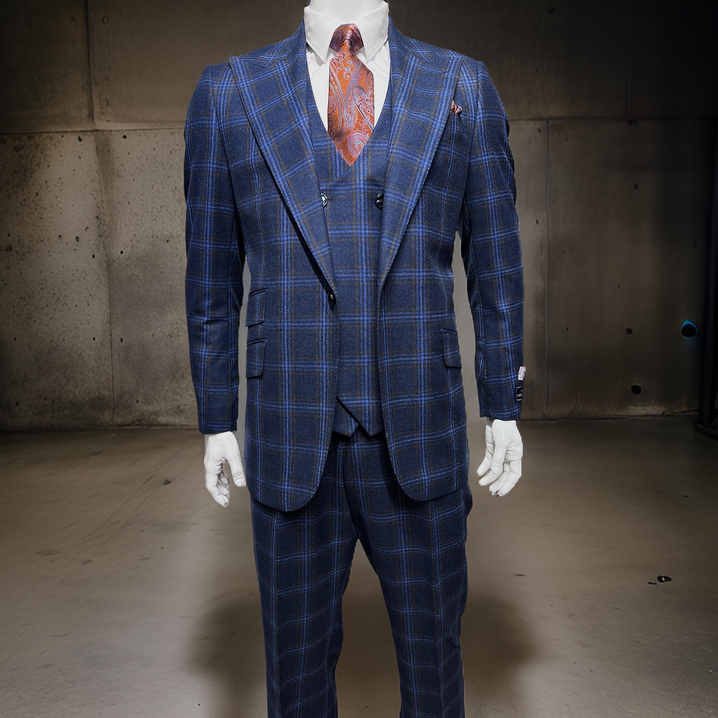 The mannequin displays a striking Tiglio Rosso Orvietto Blue/Rust Windowpane suit (sizes 50R and 52R), made from 100% wool, with a matching waistcoat and an orange tie beautifully contrasting the dark industrial background.