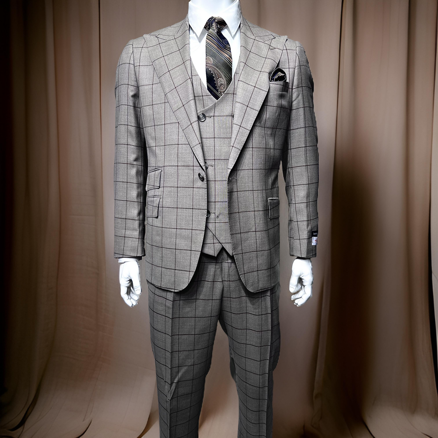 A mannequin wears a Tiglio Rosso Orvietto Grey/Brown Windowpane TL5167 suit (46R & 46L only) with a patterned tie and pocket square, set against a neutral curtain backdrop.