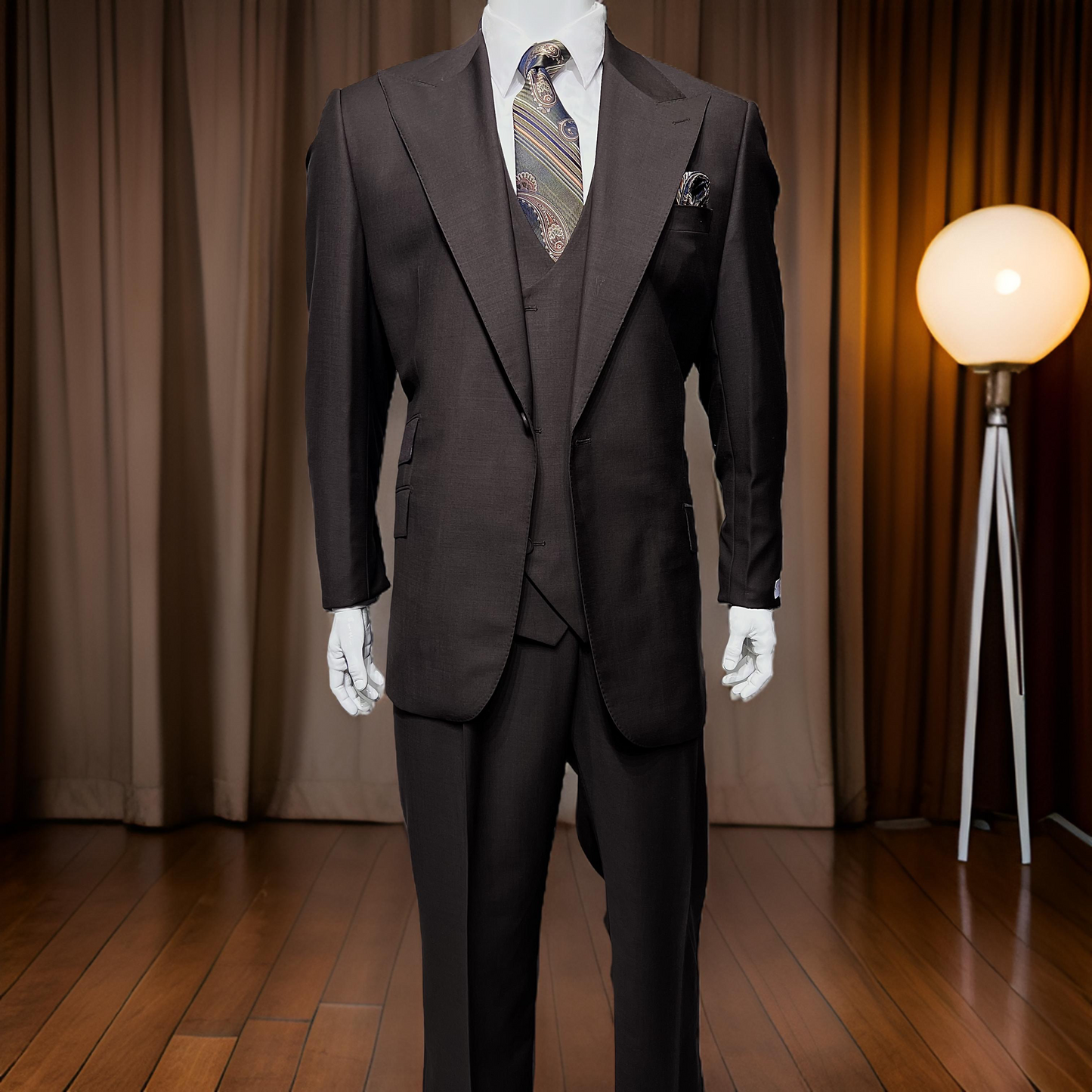 A mannequin dressed in a Tiglio Rosso Orvietto Solid Brown suit with a paisley tie stands on a wooden floor, complemented by brown curtains and a round floor lamp in the background. Expertly crafted from 100% wool, this single-pleat suit exemplifies the elegance of custom-made garments.