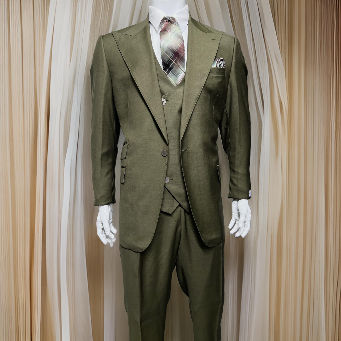 A mannequin displays the Tiglio Rosso Orvietto Solid Olive Single Pleat suit, an elegant olive green three-piece ensemble crafted from 100% wool, complemented by a plaid tie and handkerchief in the pocket, standing gracefully against a curtain backdrop.